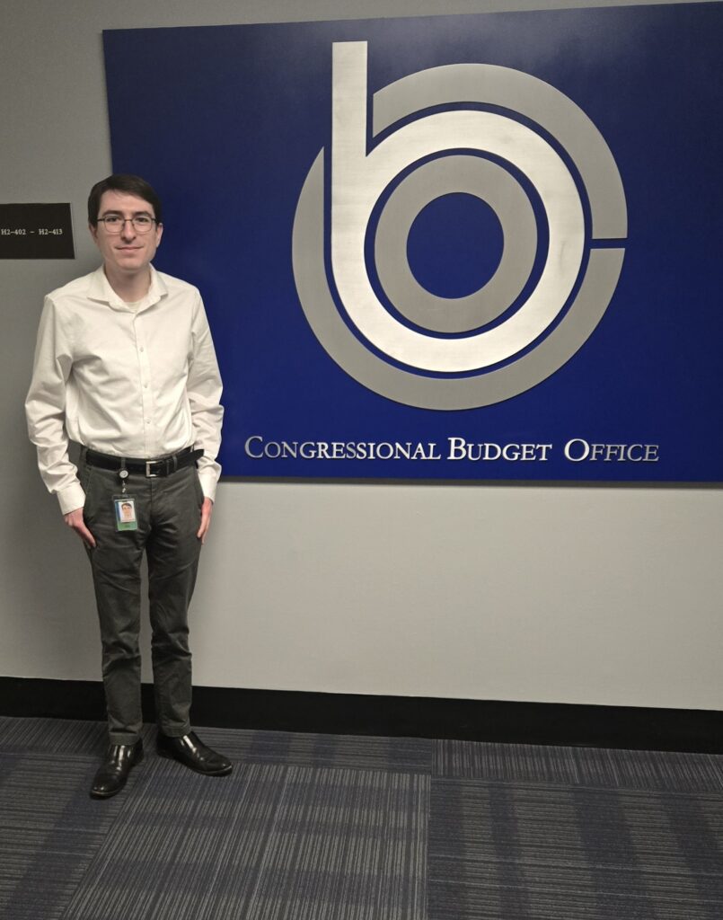 A white man with dark hair and glasses wearing a white dress shirt and dark pants next to a blue sign with a large logo in the center with the text Congressional Budget Office at the bottom.